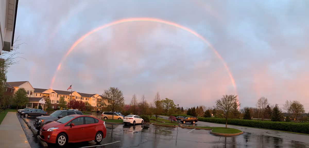 A parking lot in front of a multi-story senior living facility building with several parked cars. A vibrant rainbow arcs across a cloudy sky above the building and surrounding trees. The ground appears wet from recent rain.