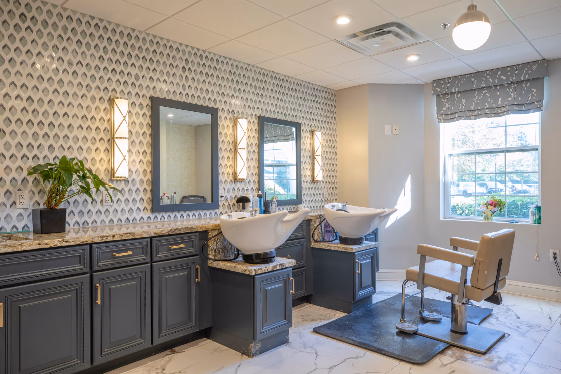 Interior of a salon area with two hair washing stations featuring white sinks and dark cabinetry. There are two mirrors on the wall with patterned wallpaper and wall-mounted lights beside each mirror. A salon chair is positioned in front of the sinks, and a window with a floral valance lets in natural light. A potted plant and some hair care products are on the countertop.
