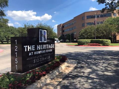 Exterior view of The Heritage at Hunters Chase assisted living and memory care facility with a large black sign displaying the facility name, address, and phone number. The building is a multi-story brick structure with windows, surrounded by trees, bushes, and a clear blue sky with some clouds.