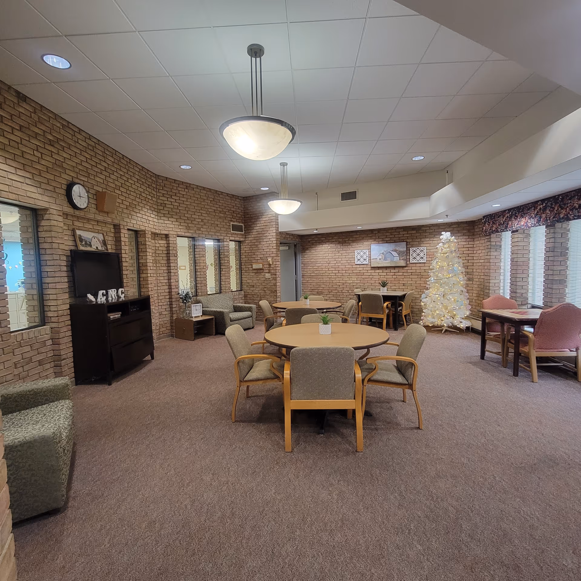 Spacious common room with round tables and chairs, brick walls, seating areas, and a decorated white Christmas tree.