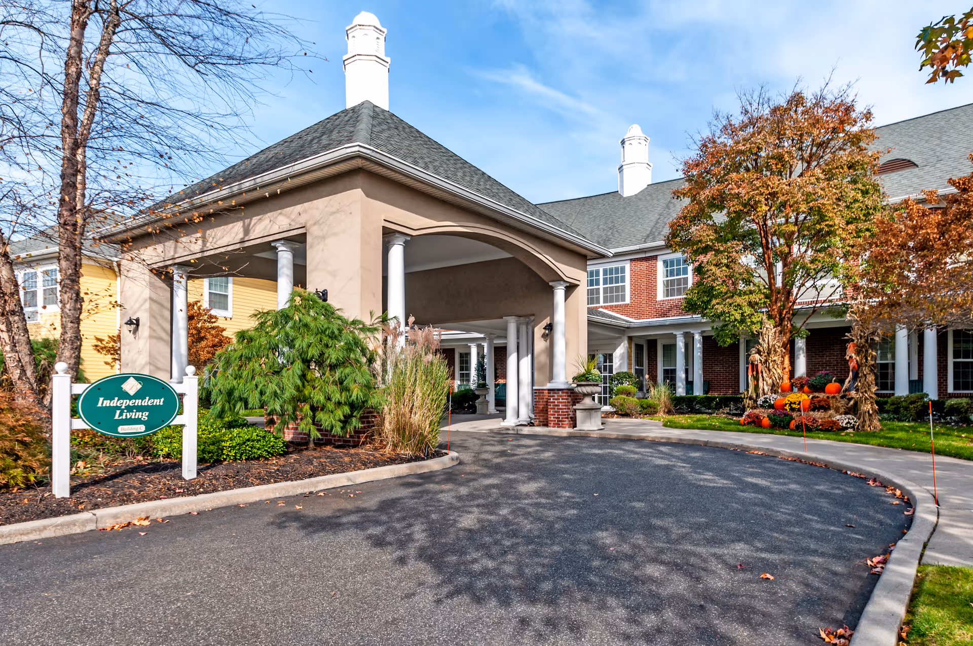Entrance to a senior living facility with a covered driveway and columns. There is a green sign that reads 'Independent Living' surrounded by landscaping with bushes and trees showing autumn colors. The building has a brick and beige exterior with white trim and multiple windows.