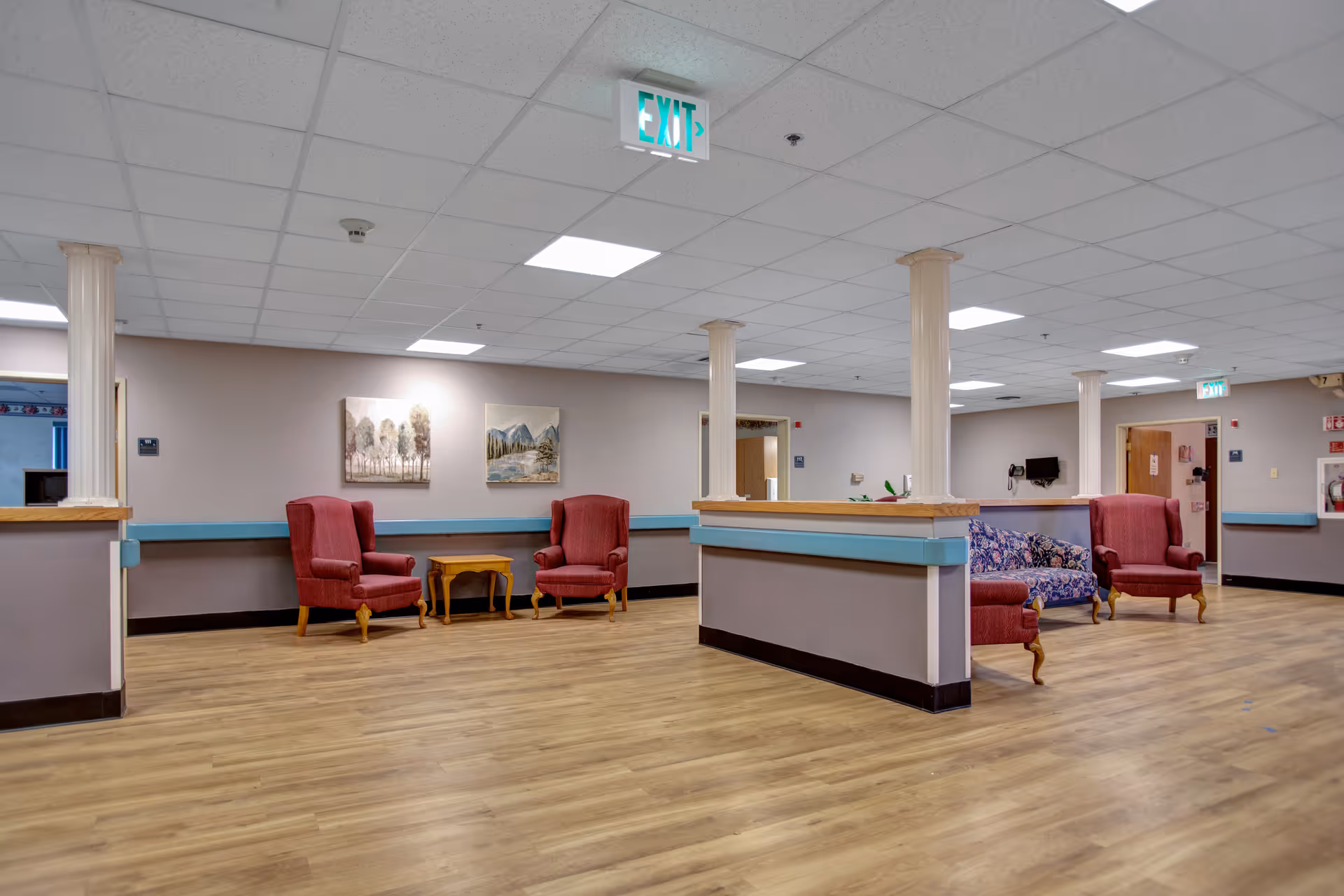 A spacious common area in a senior living facility with wooden flooring, beige walls, and a drop ceiling with fluorescent lighting. The room features several red upholstered armchairs and a floral-patterned sofa arranged around small wooden tables. There are decorative paintings on the walls and an illuminated exit sign on the ceiling. The area is divided by half walls with blue trim and supported by white columns.