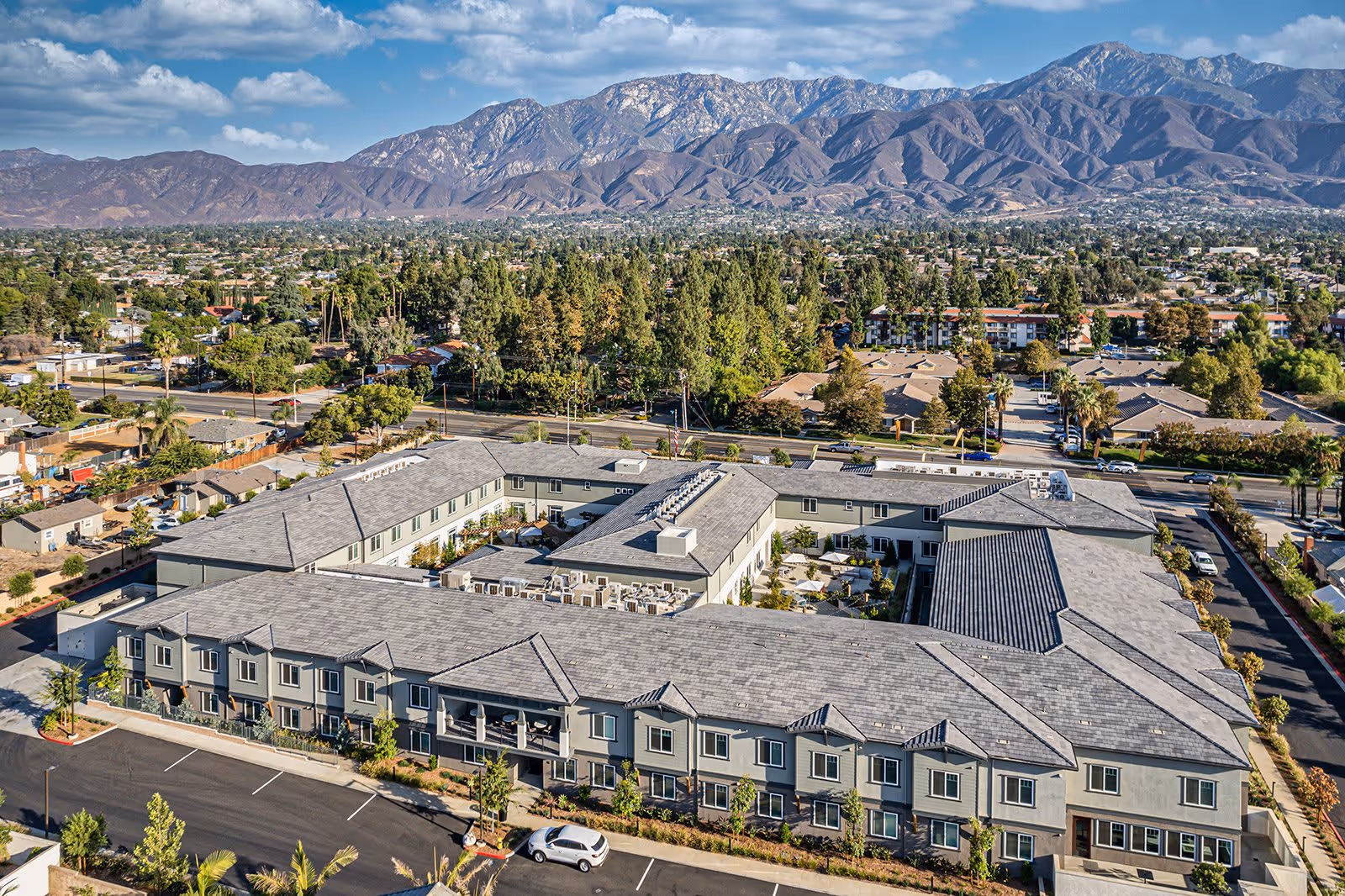 Aerial view of Allara Senior Living facility showing a large, two-story building with a gray roof and multiple windows, surrounded by trees and parking spaces. In the background, there is a sprawling suburban area with many trees and houses, and a mountain range under a partly cloudy blue sky.