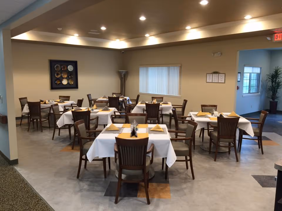 A dining room with several square tables covered with white tablecloths and yellow napkins folded on each place setting. Wooden chairs with armrests surround the tables. The room has beige walls, a window with vertical blinds, a decorative wall hanging, and a tall floor lamp in the corner. The ceiling has recessed lighting and there is an exit sign visible in the background.