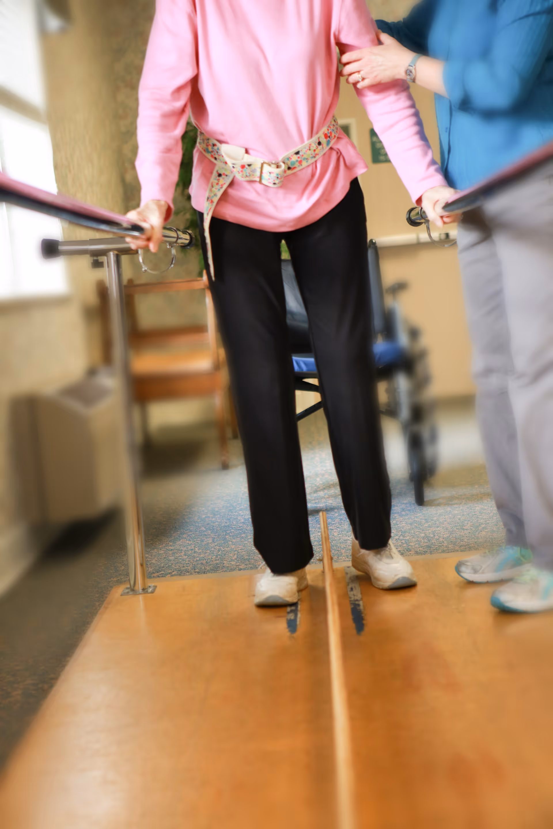 An elderly person wearing a pink top and black pants is walking with the support of parallel bars while being assisted by a caregiver dressed in blue. The elderly person is wearing a gait belt for safety during physical therapy or rehabilitation in an indoor setting.
