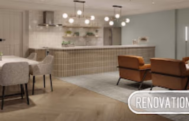 Modern open kitchen and living area with a large island, pendant lighting, beige and brown chairs, and light wood flooring in a senior living facility.
