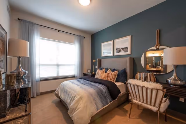 A cozy bedroom with a large window covered by sheer curtains, a bed with multiple pillows and a blue and white comforter, a dark teal accent wall with two framed pictures, a round mirror hanging above a desk with books and a lamp, and a chair with a furry cushion.