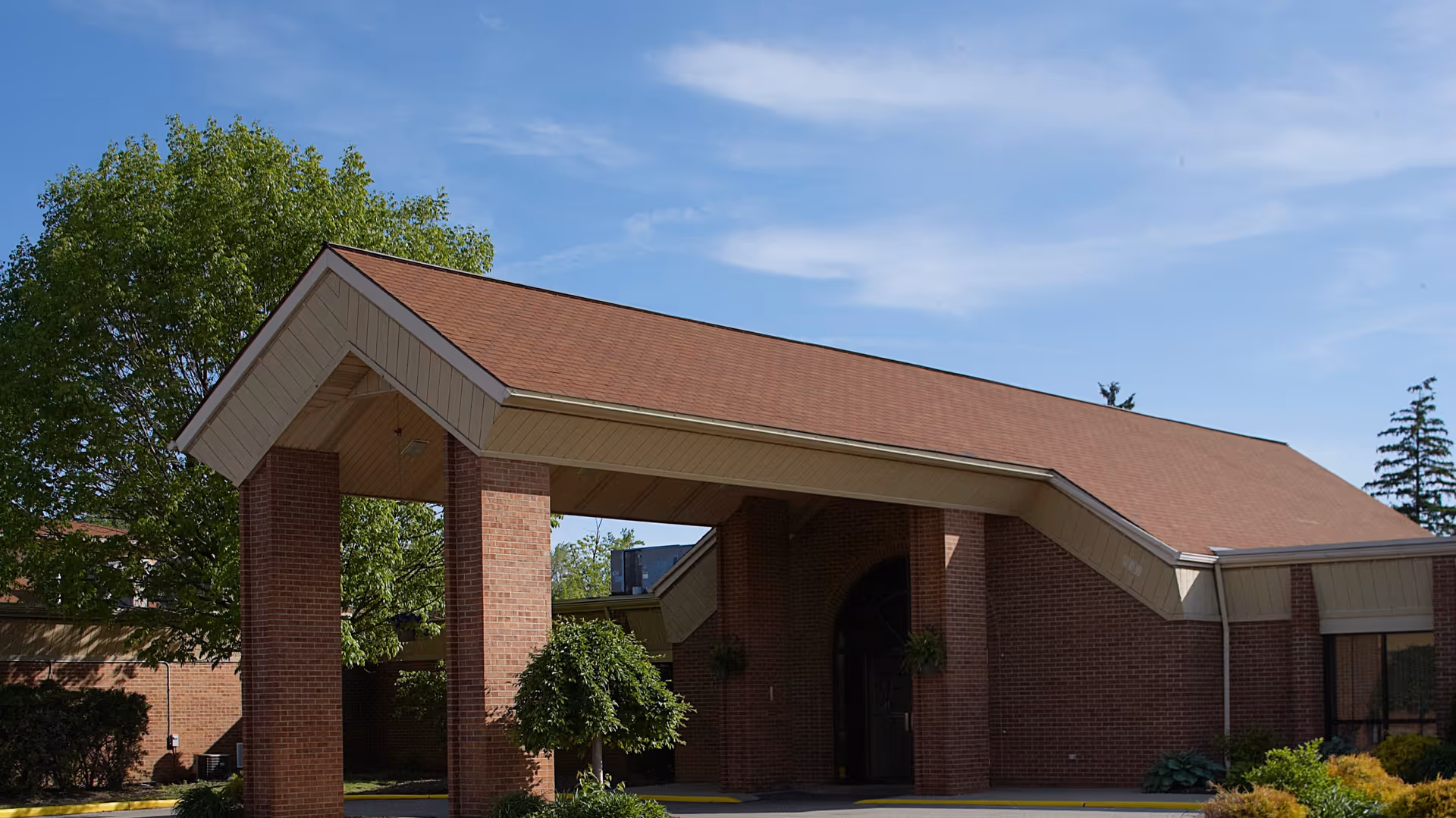 Exterior view of a brick building with a covered entrance supported by four brick pillars, surrounded by greenery and trees under a partly cloudy blue sky.