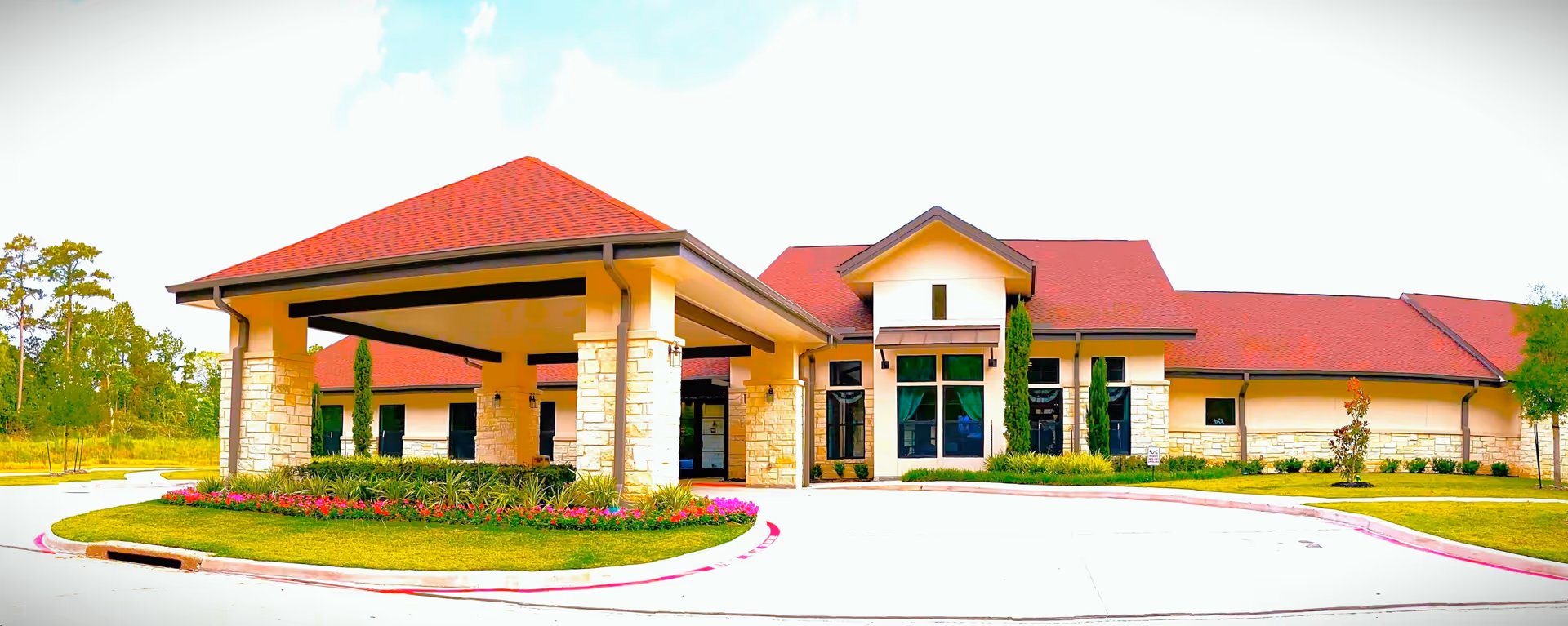 Front exterior of a single-story senior living facility with a covered porte-cochere, landscaped circular drive, and red roofs.