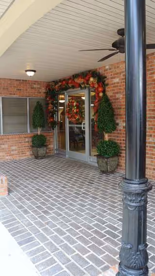 Covered brick entrance with wreaths, potted evergreens, and glass doors.