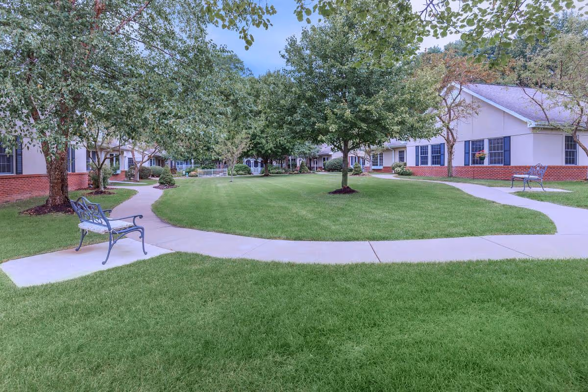 A well-maintained outdoor courtyard area with green grass, several trees, and paved walkways. There are benches placed along the paths, and single-story buildings with white walls and red brick bases surround the courtyard.