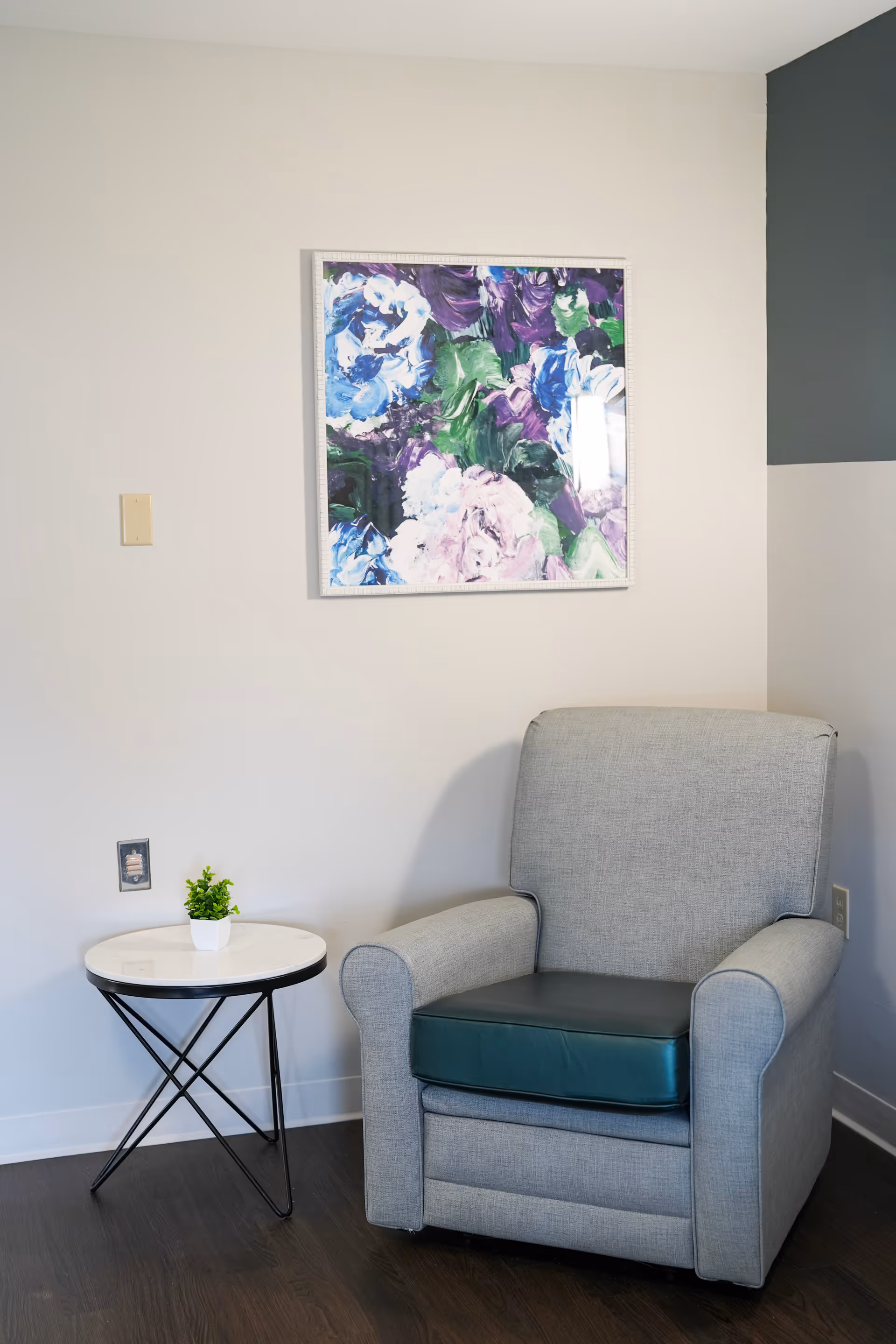 A cozy corner with a gray upholstered armchair featuring a dark green seat cushion, next to a small round white table with black metal legs holding a small potted plant. Above the chair is a colorful abstract floral painting on a light-colored wall.