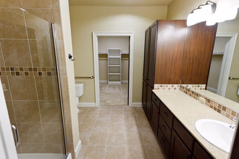 A bathroom with beige tiled floor and walls, featuring a glass-enclosed shower on the left, a white toilet next to it, and a large wooden vanity with a sink and mirror on the right. In the background, there is an open doorway leading to a walk-in closet with carpeted flooring and built-in shelving.