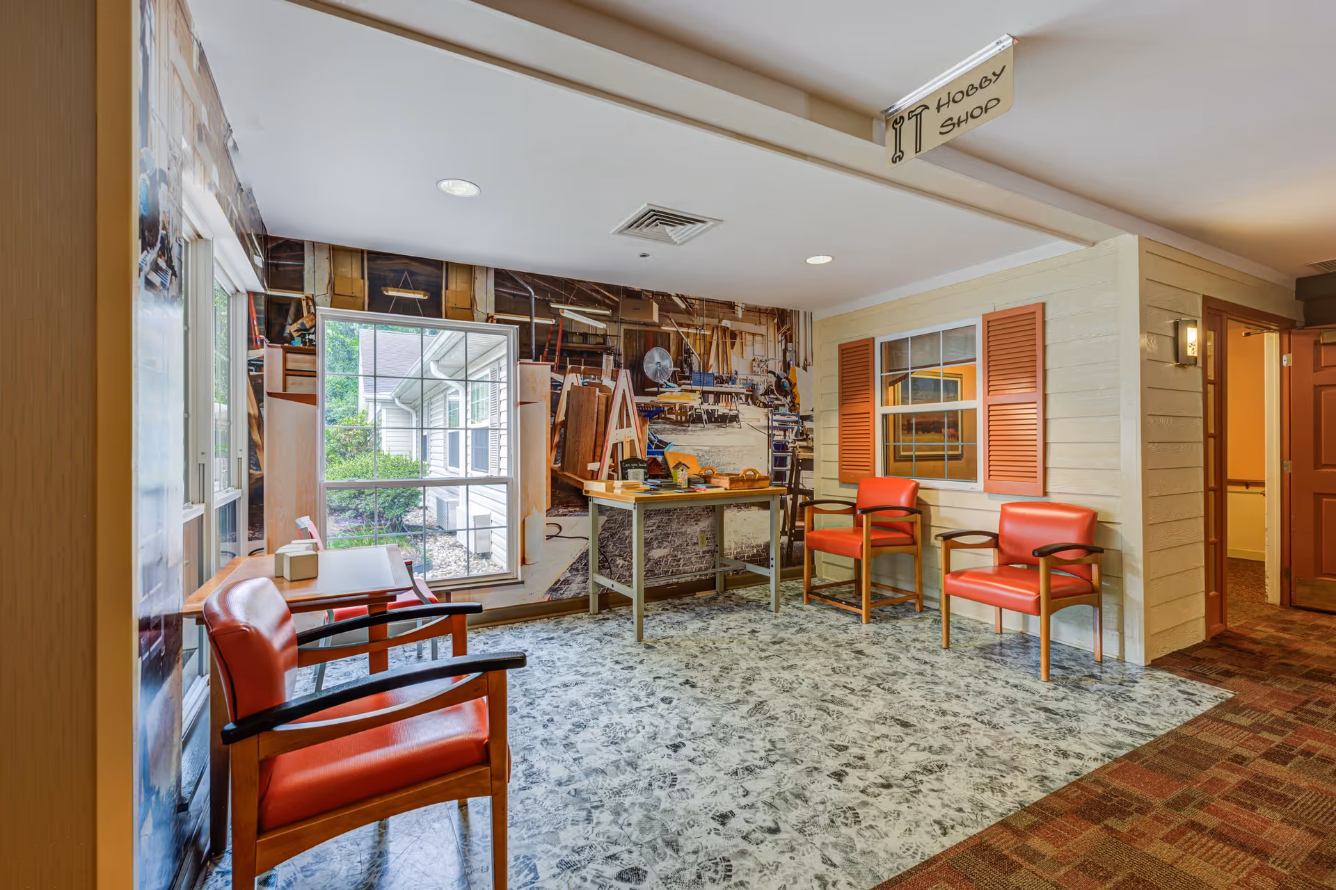 A hobby shop area inside Arden Courts - ProMedica Memory Care Community (Fair Oaks) featuring a small table with woodworking models and tools, surrounded by four red cushioned chairs with wooden arms. The walls have a large mural depicting a workshop and a window with orange shutters. The floor has a patterned carpet and linoleum. A sign hanging from the ceiling reads 'Hobby Shop'.