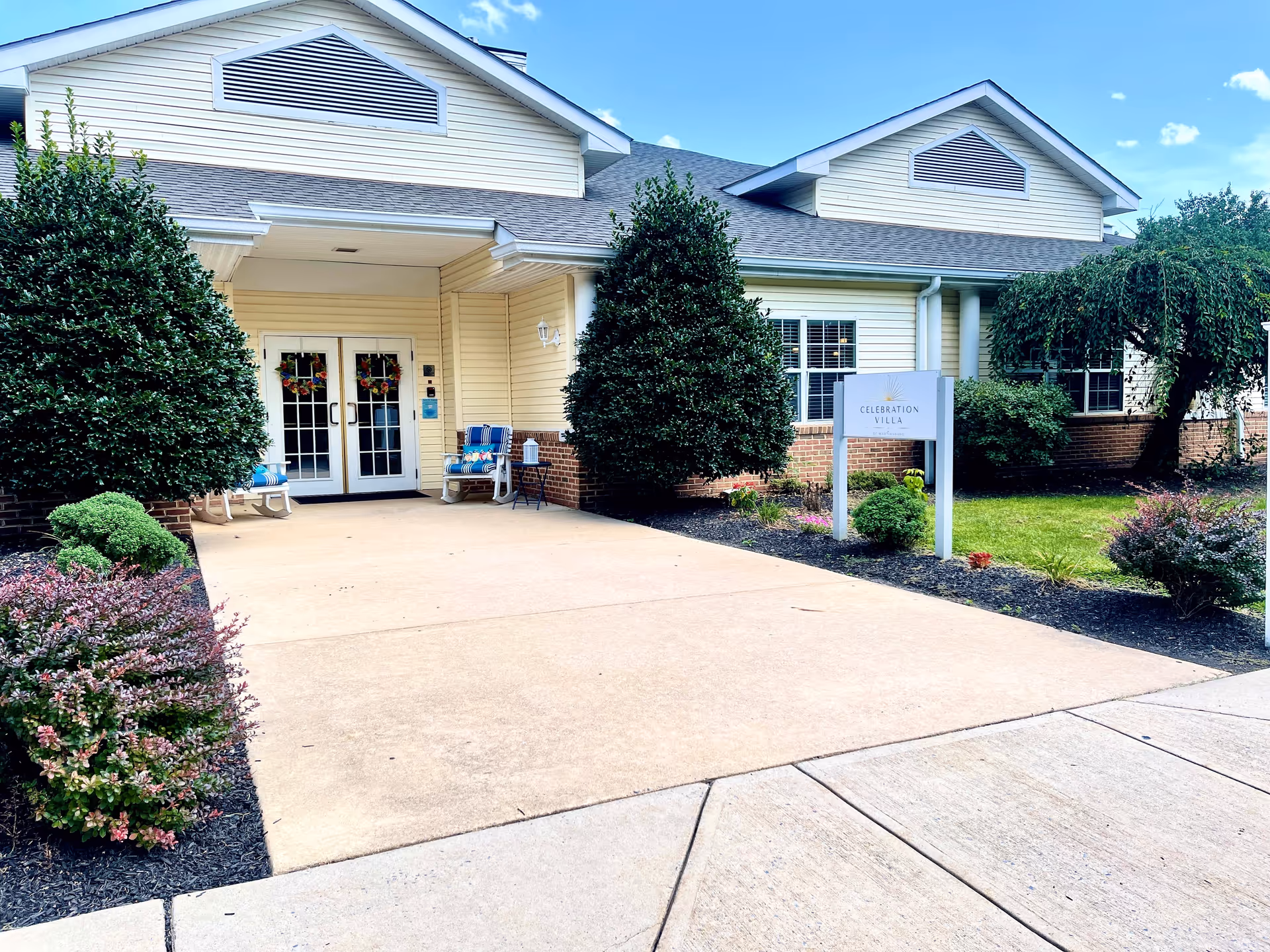 Front entrance of Celebration Villa of Martinsburg, showing a paved walkway leading to double glass doors decorated with wreaths. The building has light yellow siding with brick accents and is surrounded by neatly trimmed bushes and landscaped greenery under a blue sky.