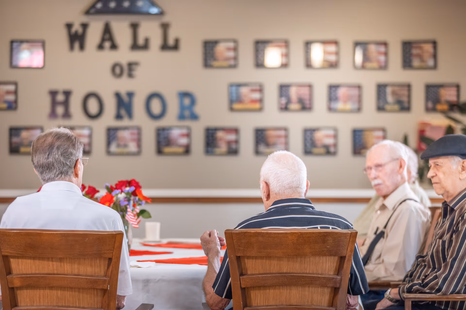 Four elderly men sitting at a table in a room with a 'Wall of Honor' display featuring framed photos and American flags on the wall behind them.