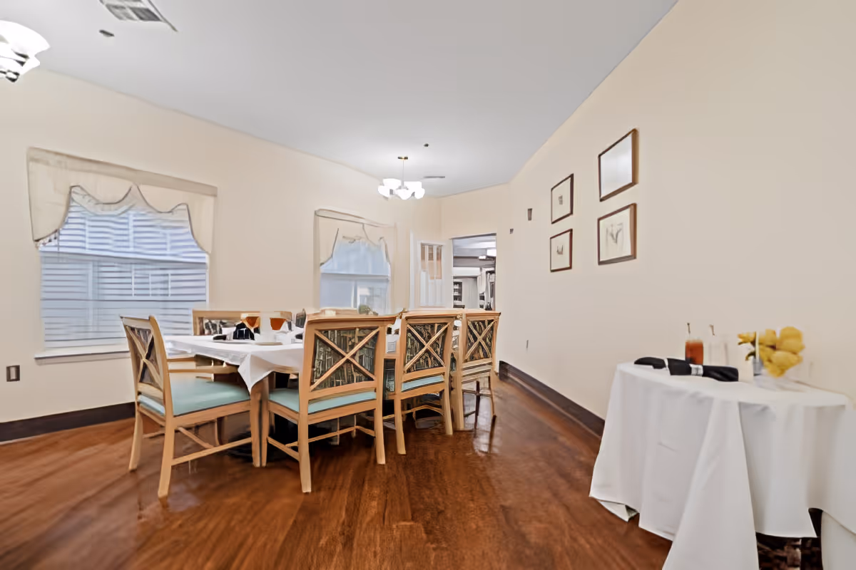 A dining room with a wooden table set for six people with chairs that have light blue cushions. The room has two windows with white blinds and beige valances. On the right side, there is a small round table covered with a white tablecloth, holding two glasses of iced tea and a vase with yellow flowers. The walls are beige with four framed pictures hanging on the right wall. The floor is wooden, and a ceiling light fixture is visible.