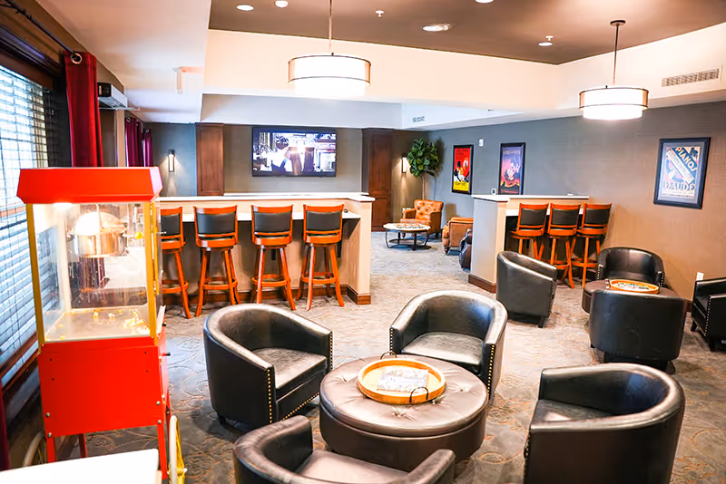 Community lounge with leather chairs and ottomans around low tables, bar stools facing a wall-mounted TV, and a popcorn machine by the window.