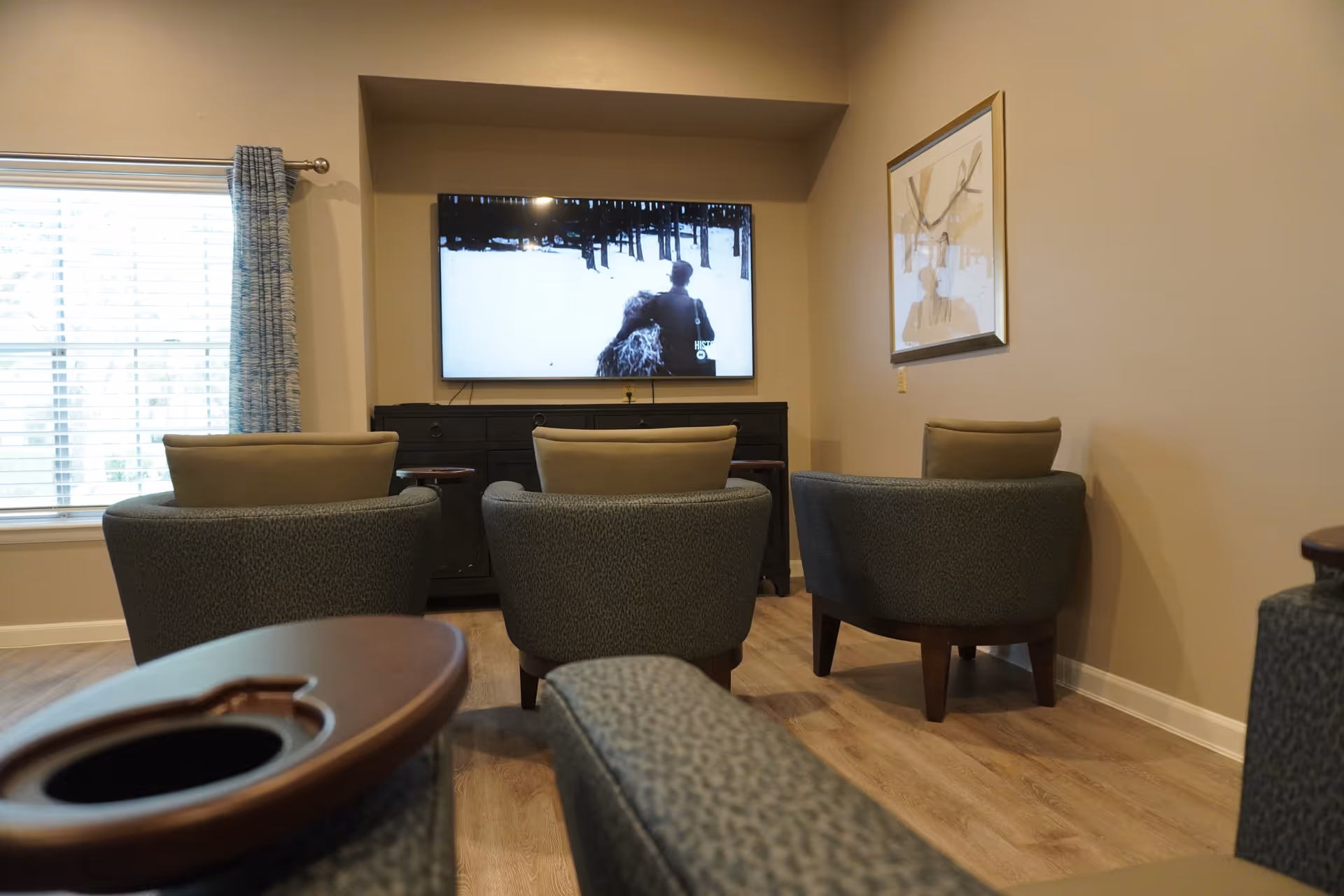 A cozy living room area with four cushioned armchairs arranged facing a wall-mounted flat-screen TV displaying a black and white scene. The room has light-colored walls, a large window with blue curtains on the left, a wooden floor, and a framed picture on the right wall.