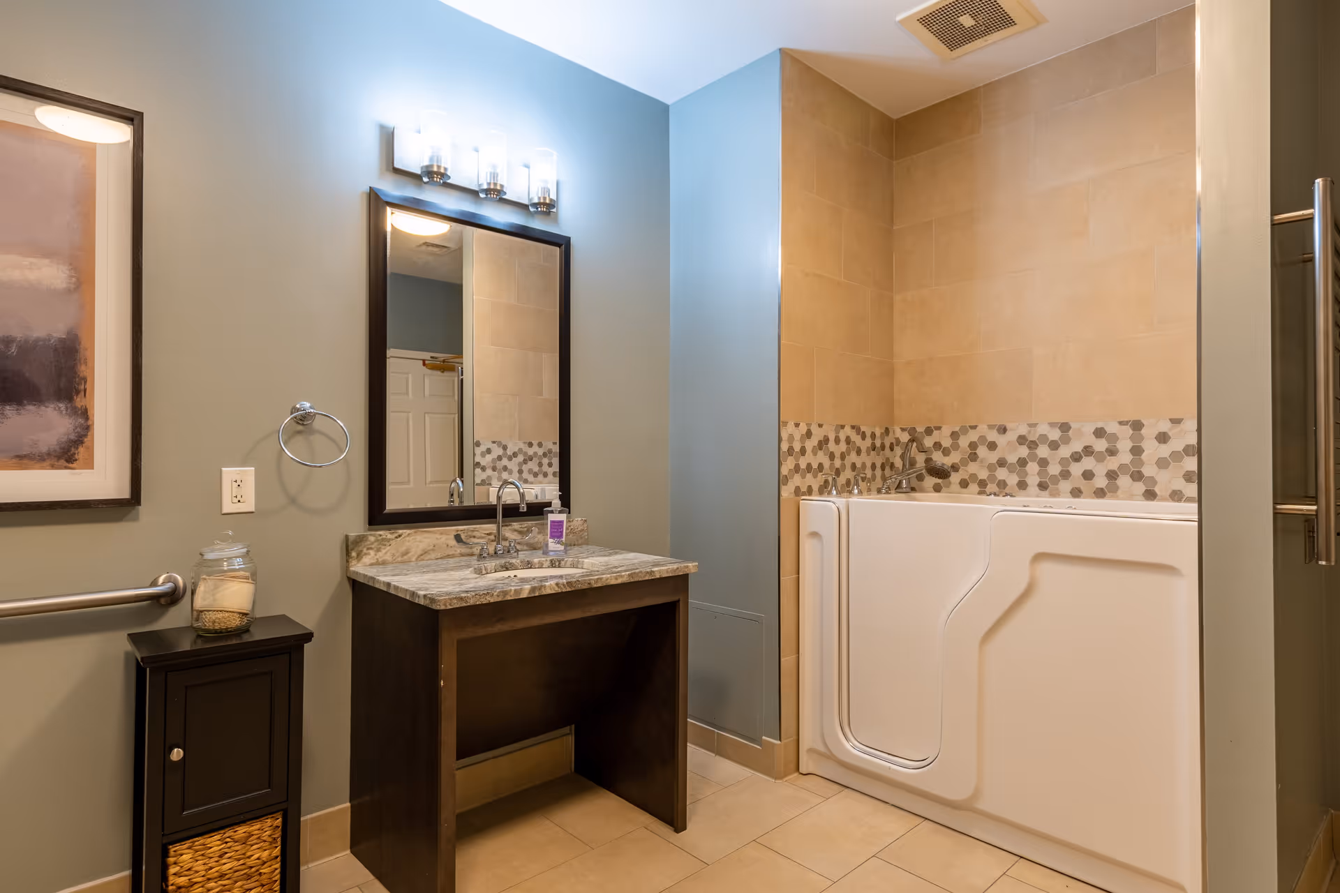 Bathroom with a walk-in bathtub featuring a side door, beige tiled walls with a decorative tile border, a dark wood vanity with a marble countertop and a sink, a large mirror with three light fixtures above it, a towel ring, a small cabinet with a glass jar on top, and a grab bar on the wall.