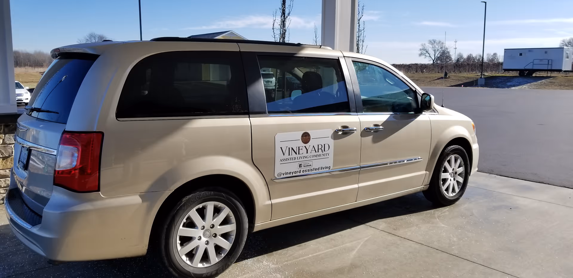 A beige minivan parked under a covered area with a sign on the side door that reads 'Vineyard Assisted Living Community'. The background shows an open paved area with some grass and a trailer in the distance under a clear blue sky.