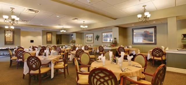 A spacious dining room with multiple round tables covered with beige tablecloths, each set with folded white napkins, silverware, and glasses. The room features wooden chairs with cushioned seats and backs, elegant chandeliers hanging from the ceiling, framed artwork on the walls, and a warm, inviting atmosphere.