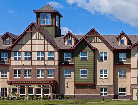 Exterior view of a multi-story senior living facility building with a combination of beige, green, and brown colors. The building features multiple windows, a tower-like structure at the center, and a well-maintained lawn with outdoor seating and umbrellas in front.