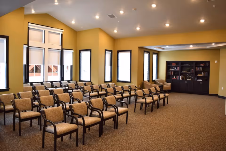 A spacious room with multiple rows of beige cushioned chairs with dark wooden arms and legs arranged facing forward. The walls are painted yellow, and there are several tall windows with white blinds allowing natural light to enter. In the back corner, there is a dark wooden bookshelf filled with books and decorative items, and two beige armchairs are positioned nearby. The ceiling has recessed lighting.