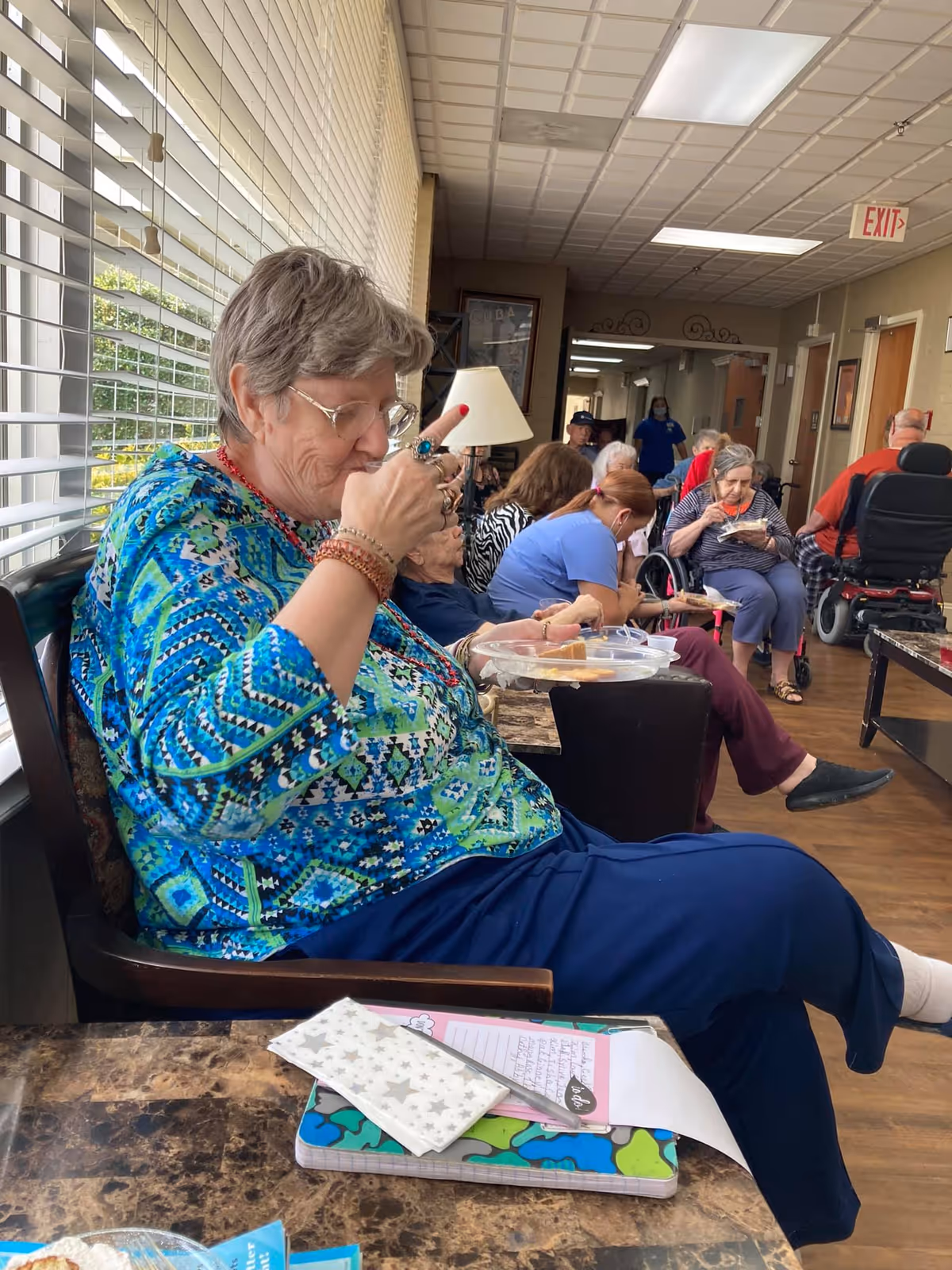 Elderly residents seated in a bright communal lounge, with a woman in a patterned shirt eating from a plate in the foreground.