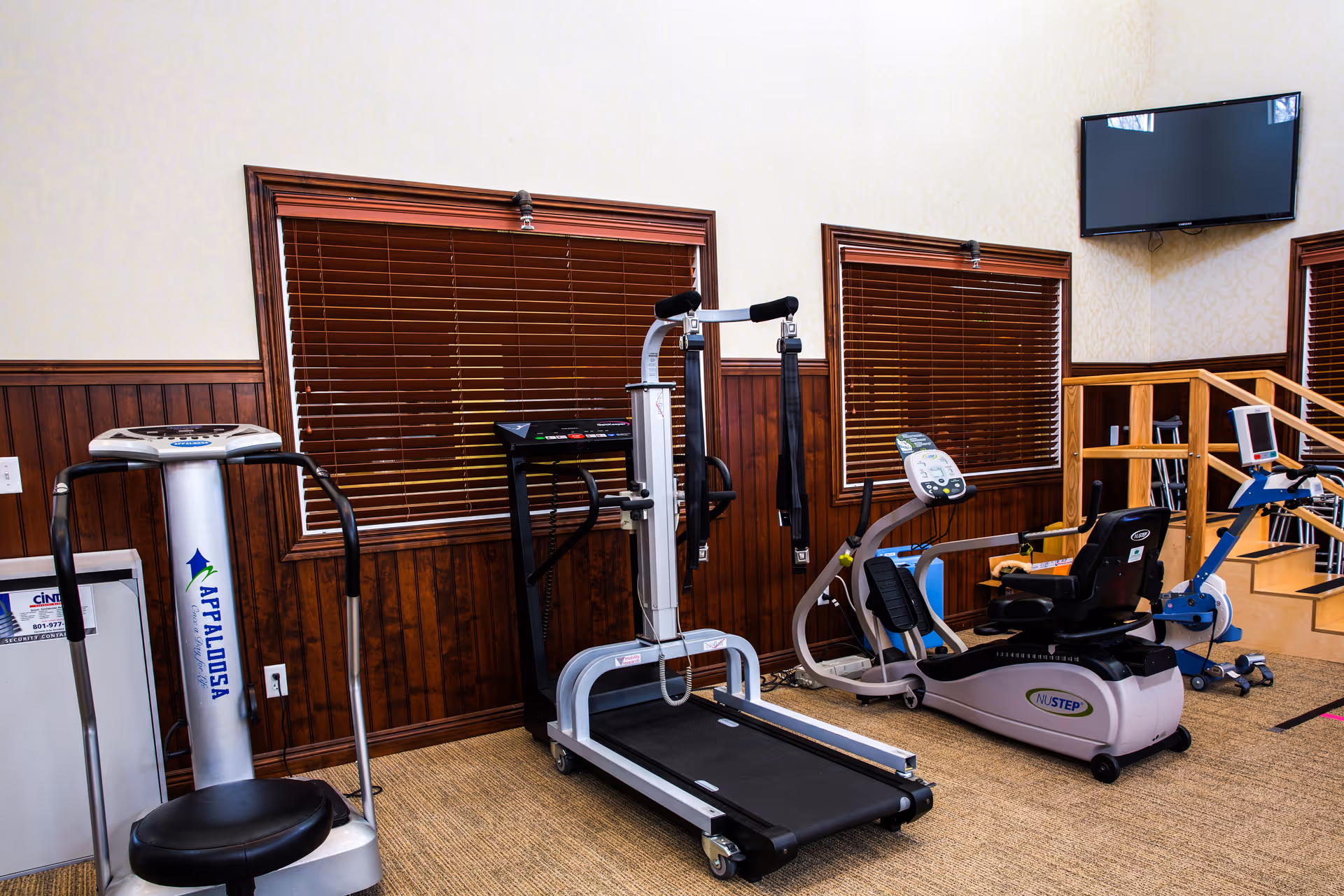 A fitness room with exercise equipment including a vibration plate machine, a treadmill, a recumbent exercise bike, and a rowing machine. The room has wood-paneled walls with two windows covered by wooden blinds and a wall-mounted TV in the corner.