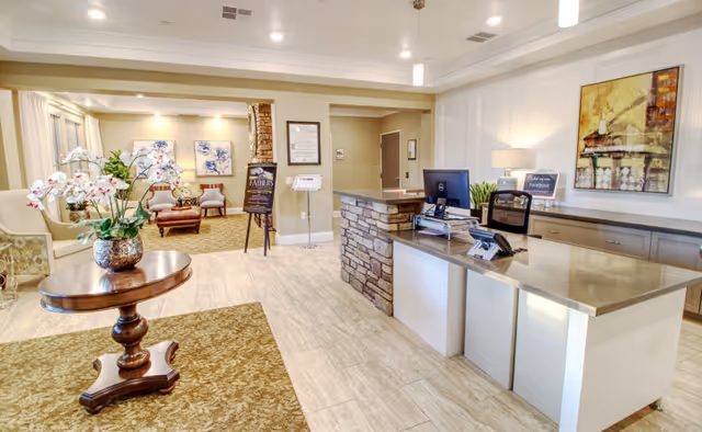 A bright and welcoming reception area in a senior living facility with a stone and wood front desk, a computer and phone on the desk, a round wooden table with a vase of white orchids, comfortable seating with chairs and a small table in the background, and framed artwork on the walls.