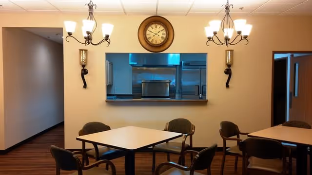 Interior view of a dining area with two square tables surrounded by chairs. The room has beige walls, wooden flooring, and two chandeliers with multiple lights hanging from the ceiling. A large round clock is mounted on the wall above a pass-through window that looks into a kitchen area with stainless steel appliances.