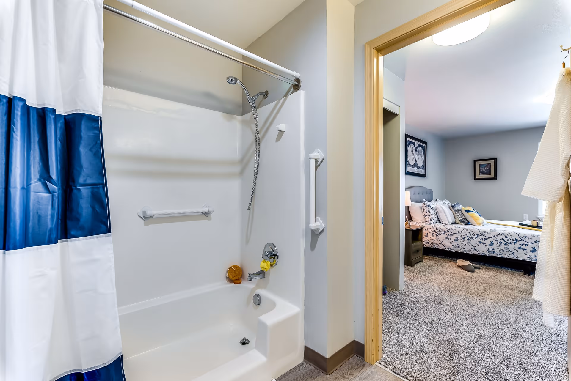 Bathroom with a white bathtub and shower combination featuring a blue and white shower curtain. The bathroom has grab bars installed on the wall. Through the doorway, a bedroom is visible with a bed covered in a floral bedspread, a nightstand with a lamp, and framed pictures on the walls.