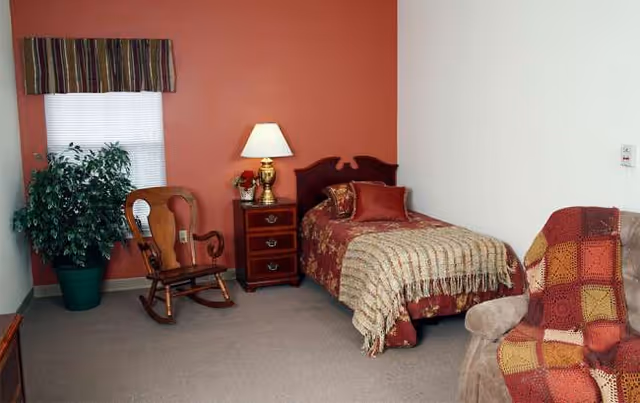 A cozy bedroom in a senior facility with a bed, rocking chair, nightstand, and plant.