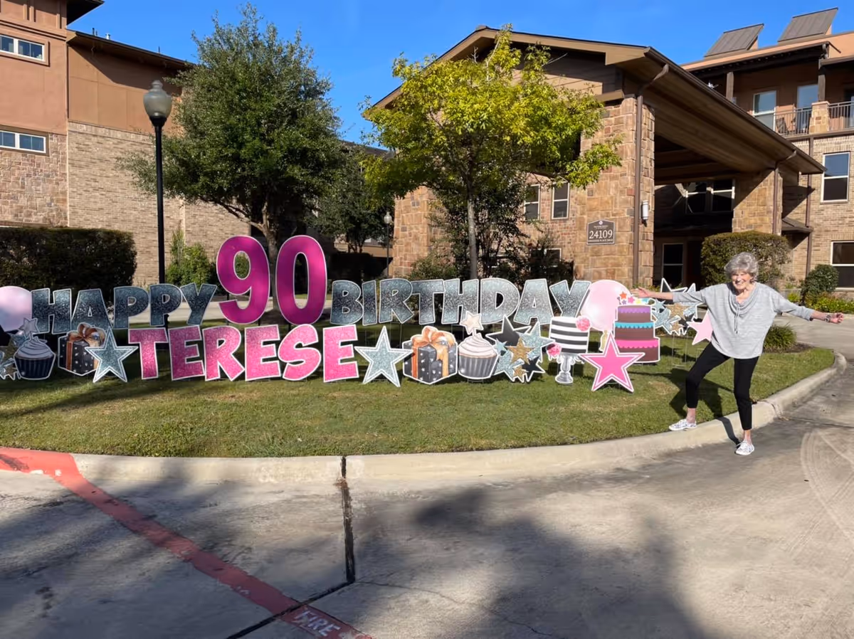 An elderly woman joyfully posing next to a large outdoor sign that reads 'Happy 90 Birthday Terese' with decorative stars, cupcakes, presents, balloons, and a birthday cake. The setting is outside a senior living facility with a stone building and trees in the background under a clear blue sky.