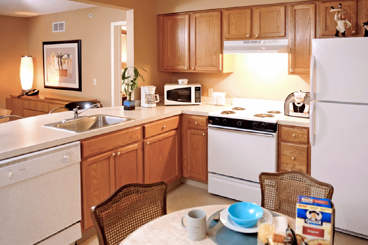 A kitchen area with wooden cabinets, a white stove, microwave, refrigerator, and dishwasher. A round dining table with two chairs is set with a blue bowl, a mug, a glass of orange juice, and a box of Quaker oatmeal. In the background, there is a living area with a lamp, framed artwork, and a plant on the counter.