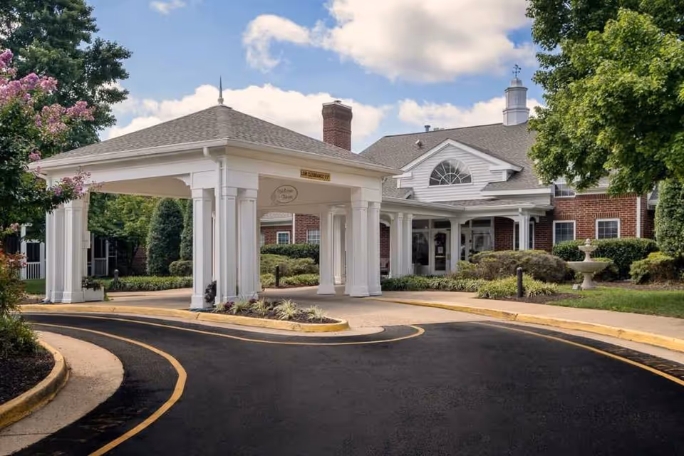 Entrance of a senior living facility named Chancellor's Village featuring a covered drop-off area with white columns, a brick building with white trim, a chimney, and landscaped greenery including trees and bushes under a partly cloudy sky.
