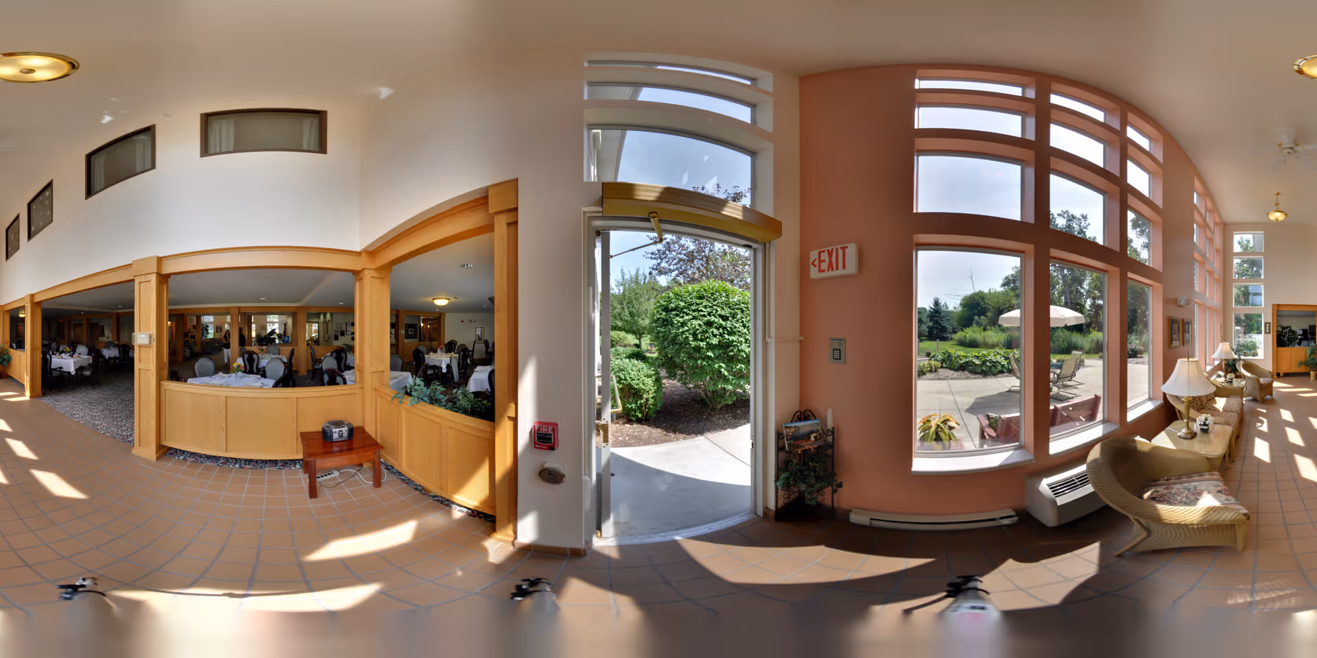 A bright interior space featuring a dining area with tables and chairs set for meals on the left, and a sunlit lounge area with wicker chairs and lamps on the right. Large windows provide a view of a patio and garden outside, with greenery and outdoor seating visible. The floor is tiled, and wooden columns and trim separate the dining area from the lounge.