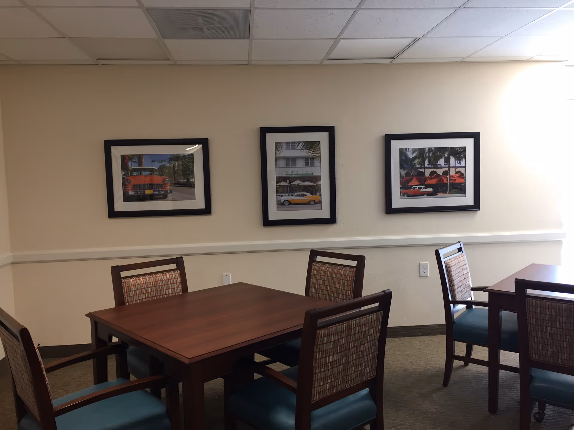 A room with two wooden tables and six chairs with patterned backs and teal seats. Three framed pictures of vintage cars and outdoor scenes hang on a beige wall. The room has a carpeted floor and a drop ceiling with fluorescent lighting.