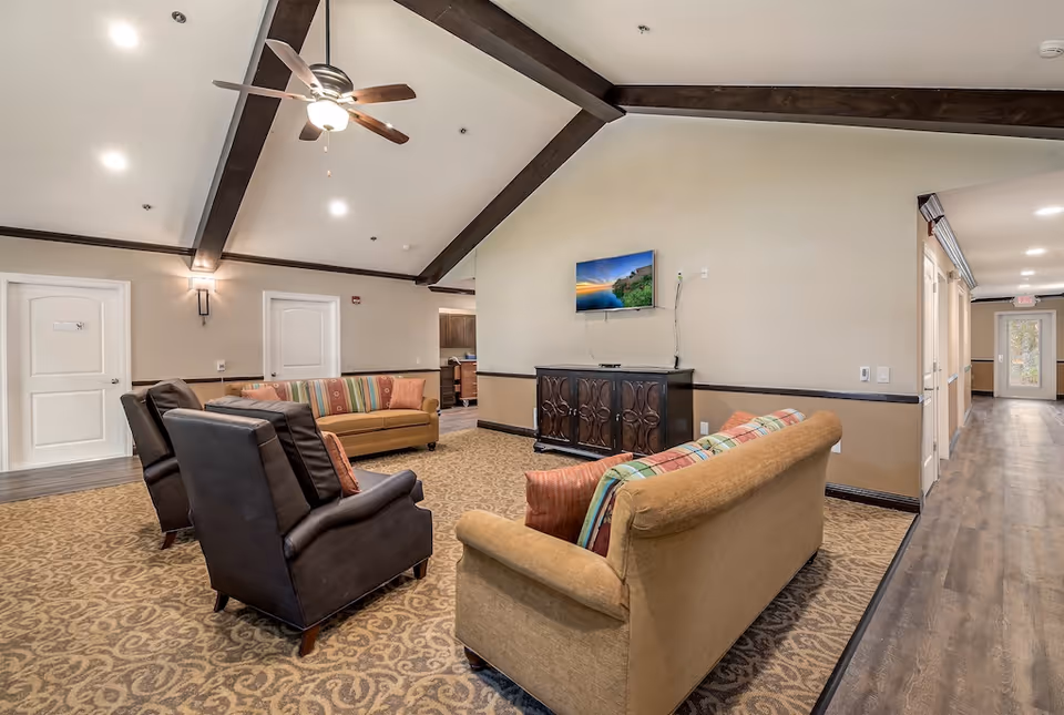 A cozy living room area in Pacifica Dogwood Cottage featuring two brown leather armchairs and two beige sofas with colorful striped cushions arranged around a dark wooden cabinet with a flat-screen TV mounted above it. The room has a high vaulted ceiling with exposed dark wooden beams and a ceiling fan with lights. The floor is carpeted with a patterned design, and there is a hallway with wood flooring leading to a door at the far end.