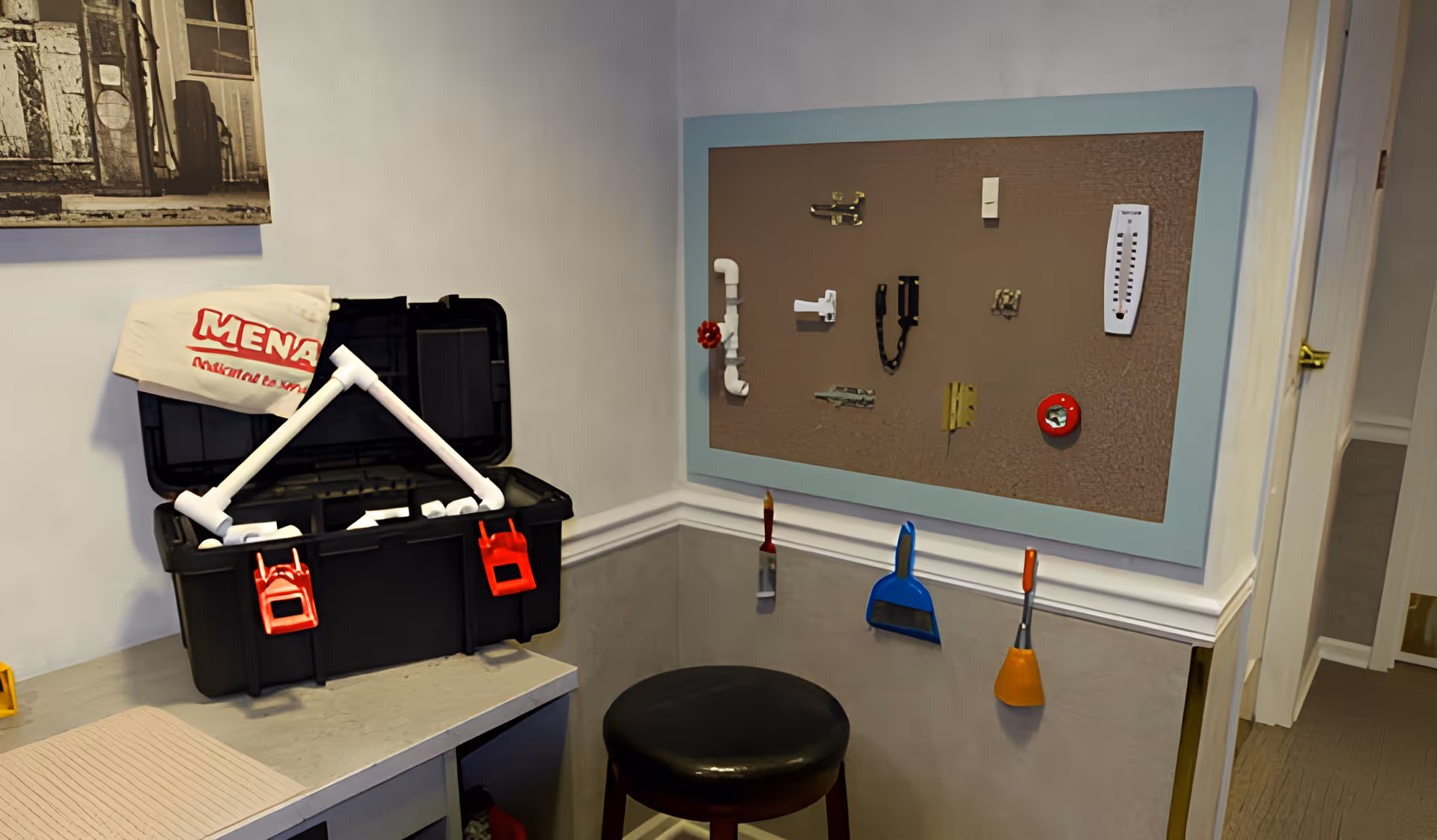 A small interior activity corner with an open black toolbox on a counter, a cork sensory board with latches on the wall, hanging cleaning tools and a black stool.