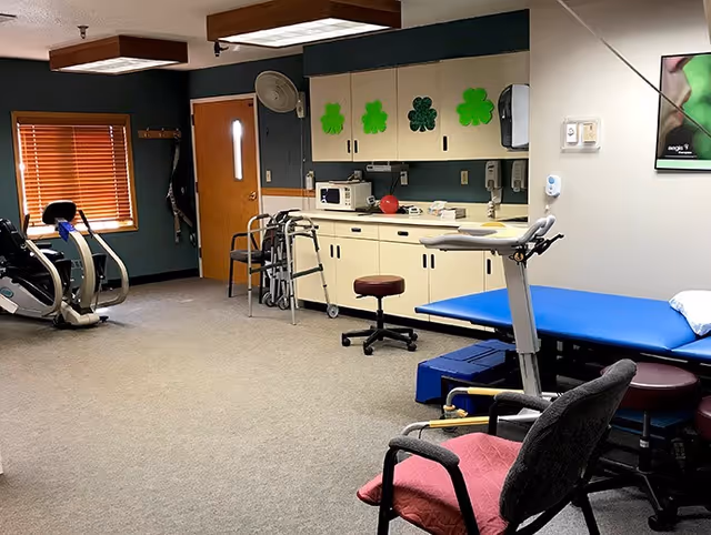 An interior therapy/exercise room with a treatment table, exercise machines, walkers, stools and cabinets decorated with shamrock cutouts.