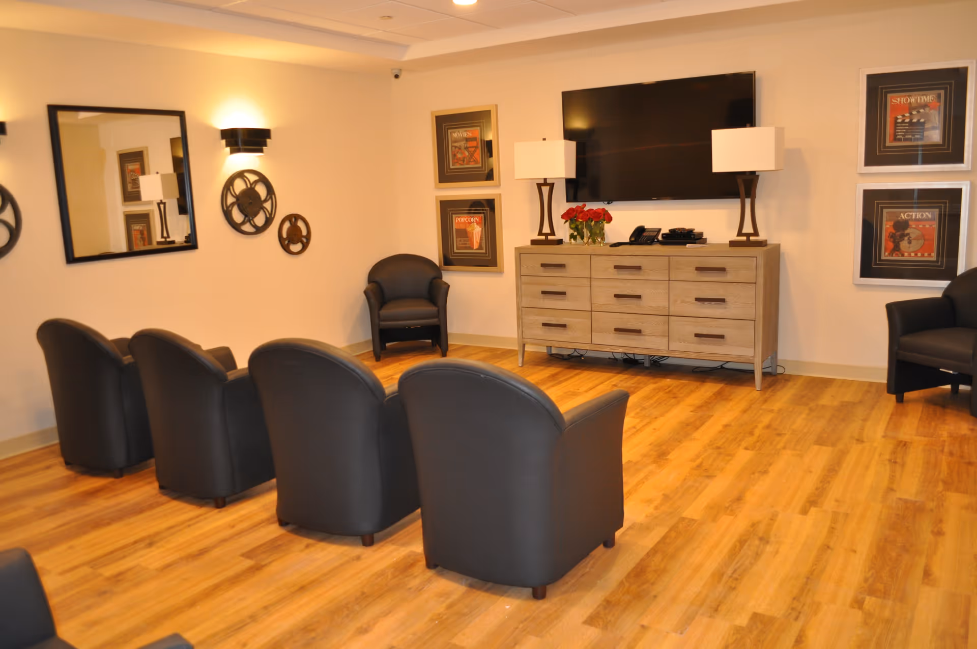 A cozy common area with four black armchairs arranged in two rows facing a large flat-screen TV mounted on the wall. Below the TV is a wooden dresser with two lamps and a vase of red flowers. The room has wooden flooring, framed movie-themed artwork on the walls, a large mirror, and decorative film reel wall art.