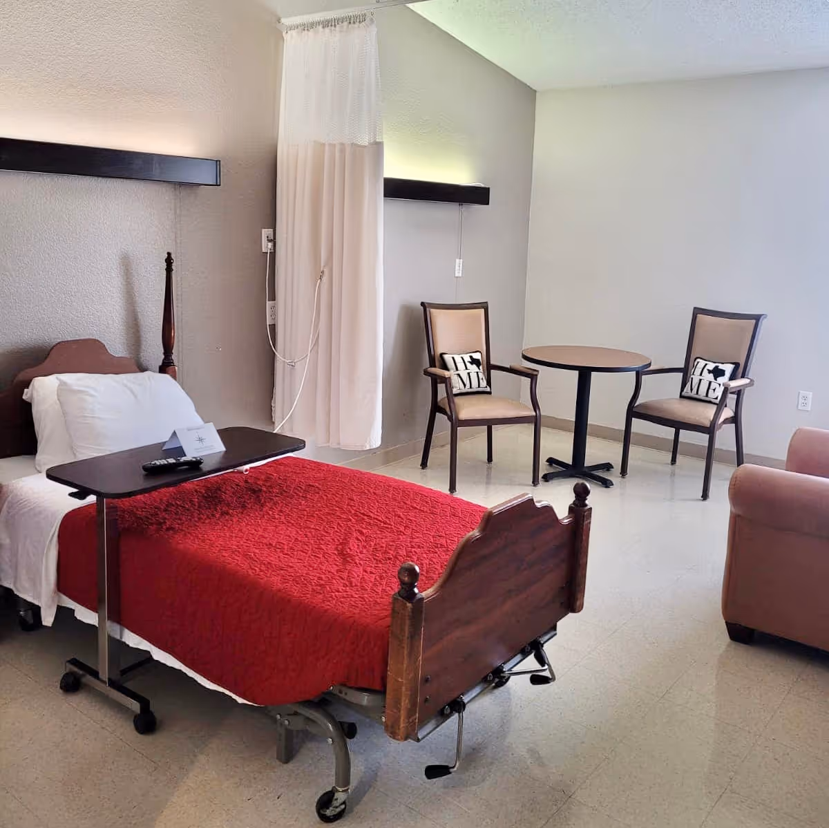 A nursing facility room with a single bed covered in a red blanket and white pillow. There is a movable overbed table with a remote control on it. Two chairs with cushions and a small round table are placed near the wall. A curtain hangs from the ceiling near the bed, and a beige armchair is partially visible on the right side.