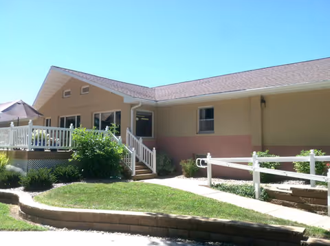 Exterior view of a single-story assisted living facility building with beige and light brown walls, a sloped roof, a white railing along a raised porch, and a wheelchair-accessible ramp surrounded by greenery and a small lawn.