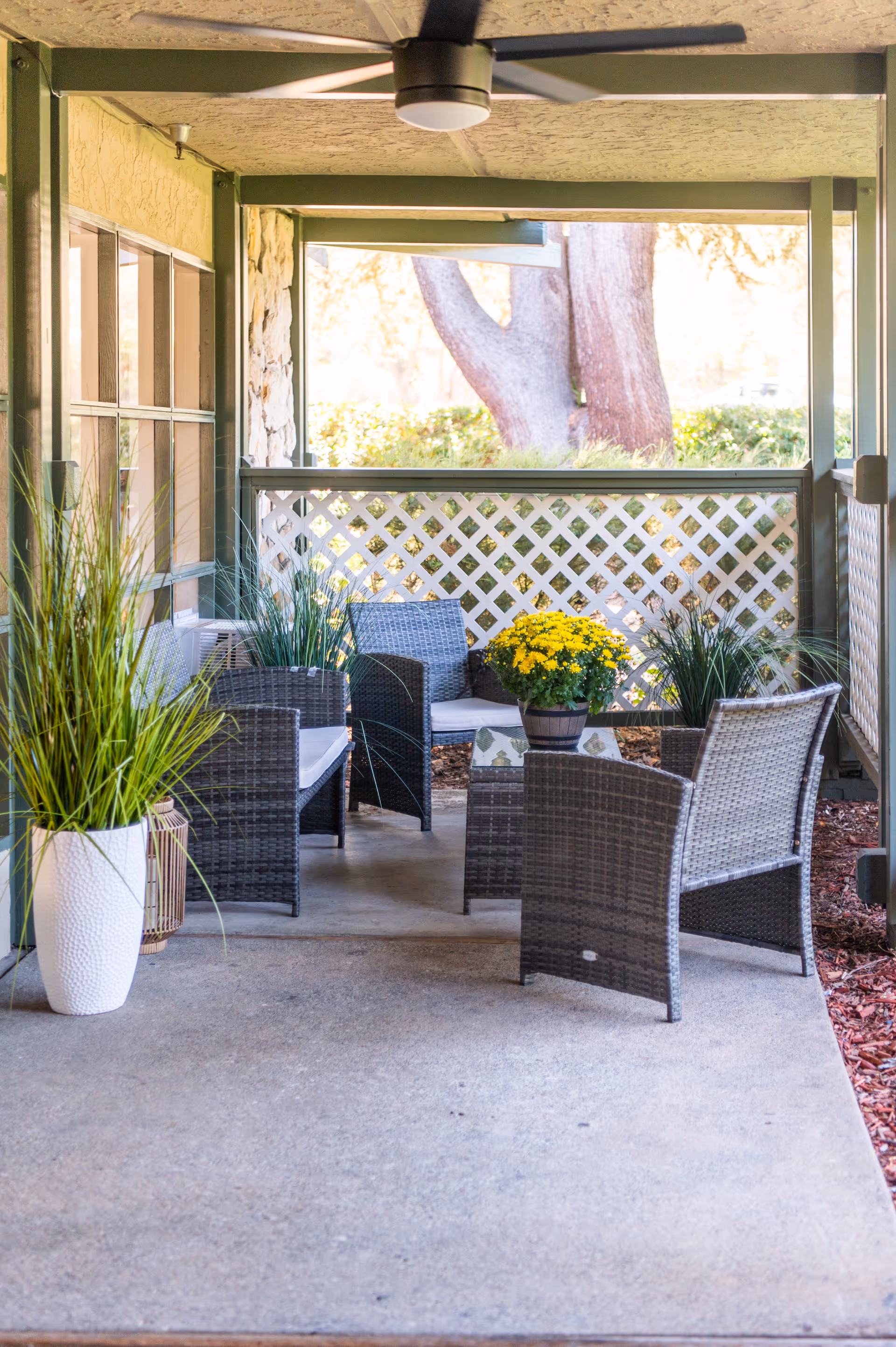 Covered outdoor patio seating area with wicker chairs and a small table, potted plants, lattice railing, and a ceiling fan.