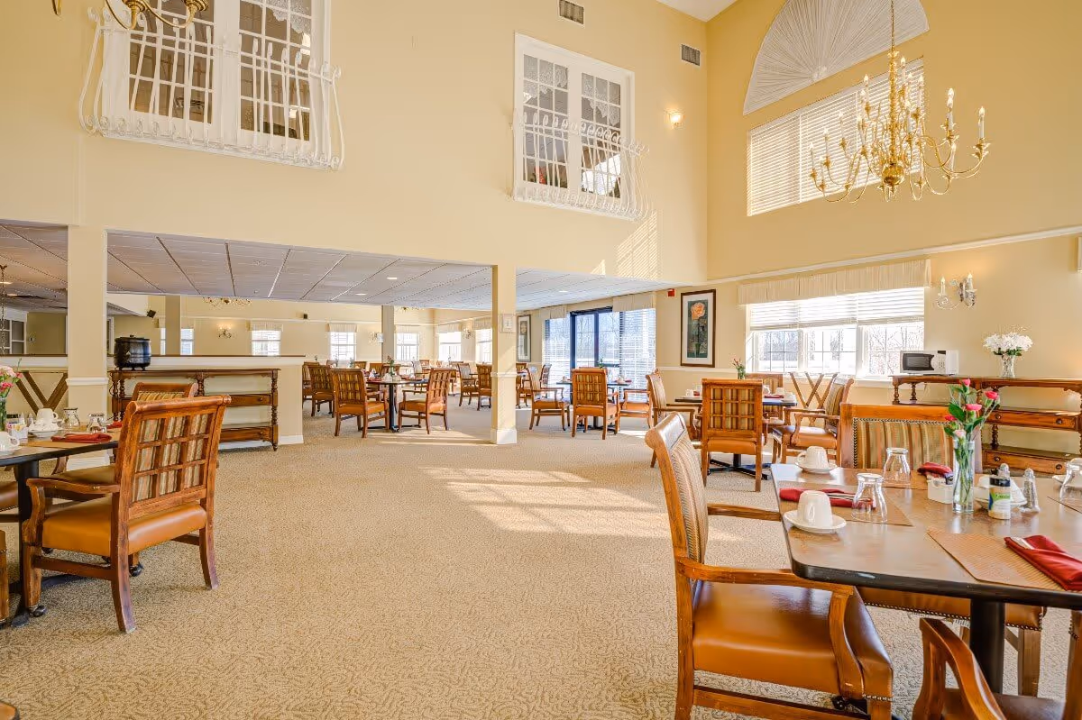 Bright and spacious dining room with multiple wooden tables and chairs arranged neatly. The room features large windows allowing natural light to fill the space, beige walls, and a high ceiling with a decorative chandelier. Tables are set with cups, glasses, napkins, and small flower vases.