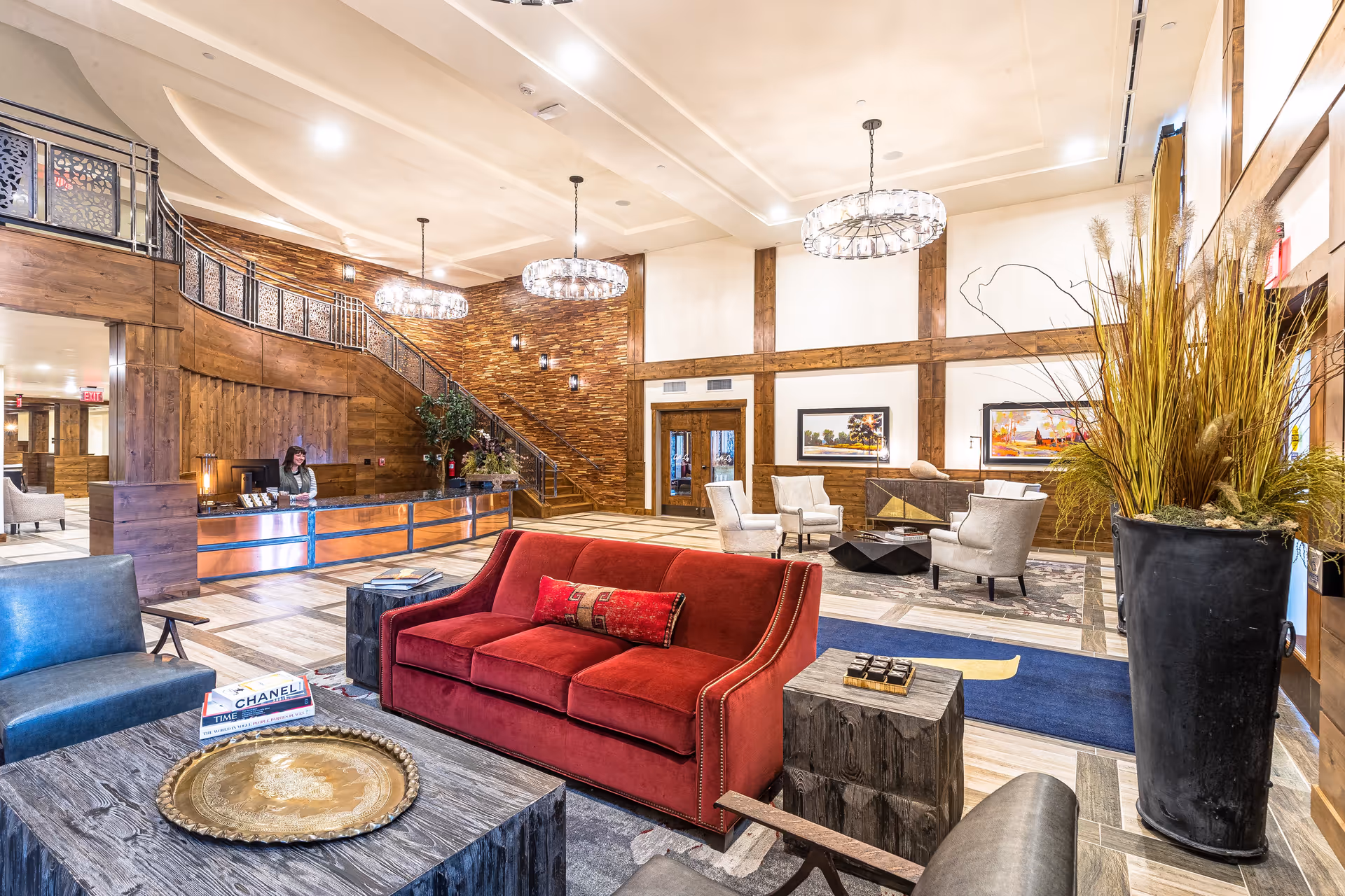 A spacious and well-lit senior living facility lobby with a red sofa, gray armchairs, wooden tables, and decorative plants. The reception desk is staffed by a woman, and there is a staircase with wooden paneling and metal railings. The walls feature wood accents and framed artwork, with modern chandeliers hanging from the ceiling.