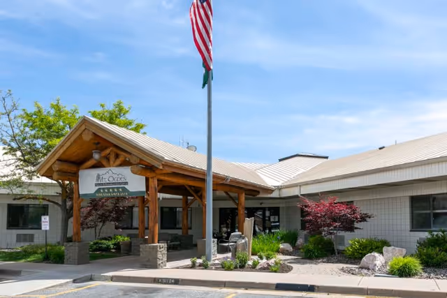 Exterior view of Mt. Ogden Health And Rehabilitation Center showing the entrance with a wooden canopy supported by stone pillars, an American flag on a flagpole in front, and landscaped greenery including bushes and small trees under a clear blue sky.