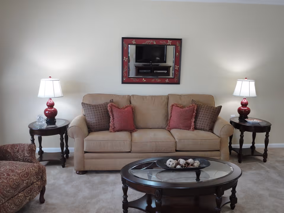A living room with a beige sofa adorned with two red and two brown patterned cushions. On either side of the sofa are dark wooden side tables, each with a red lamp with a white lampshade. A round glass coffee table with a dark wooden frame is in front of the sofa, holding a decorative bowl with assorted natural elements. A patterned armchair is partially visible on the left side. A wall-mounted mirror with a red and gold frame hangs above the sofa, reflecting a television and media console.